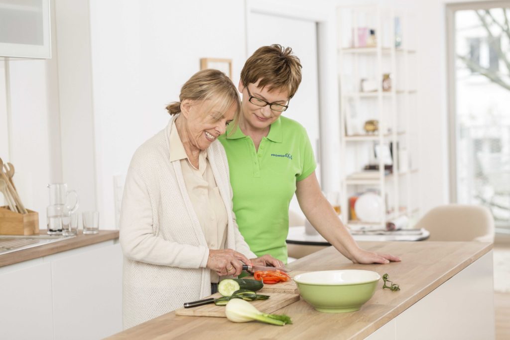 Betreuungskraft hilft Senioren in der Küche beim Gemüseschneiden.