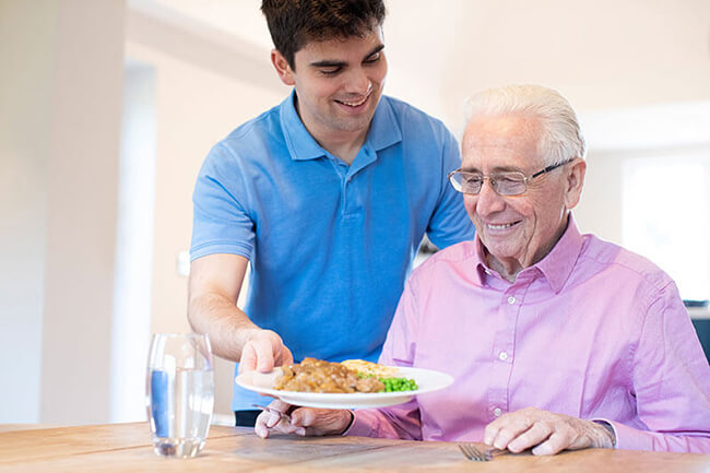 Ein Alltagsbegleiter stellt eine warme Mahlzeit auf den Tisch vor einen Senioren