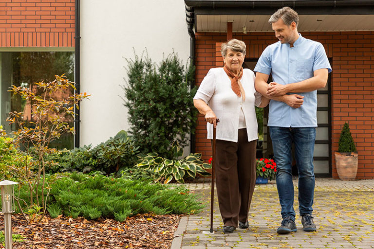 Ein Betreuer stützt eine Seniorin beim Spaziergang auf dem eigenen Grundstück